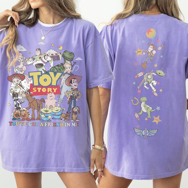 Op de afbeelding: Een lichtpaars t-shirt met Toy Story-personages op de voorkant, waaronder Woody, Buzz Lightyear en Jessie, met de tekst Toy Story en You've Got a Friend in Me. De achterkant toont een verticale rangschikking van personage-iconen.