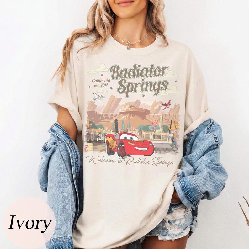 May include: Ivory t-shirt with a graphic of Radiator Springs, California, featuring the text "Radiator Springs" and "Welcome to Radiator Springs." The design includes a red car and a desert landscape.