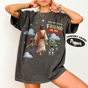May include: Dark grey t-shirt featuring the text "YOU'VE GOT A FRIEND IN ME" above an illustration of Jesus, Woody, and Buzz Lightyear. The shirt also has the words "COMFORT COLORS" and "Pepper" in a white oval.