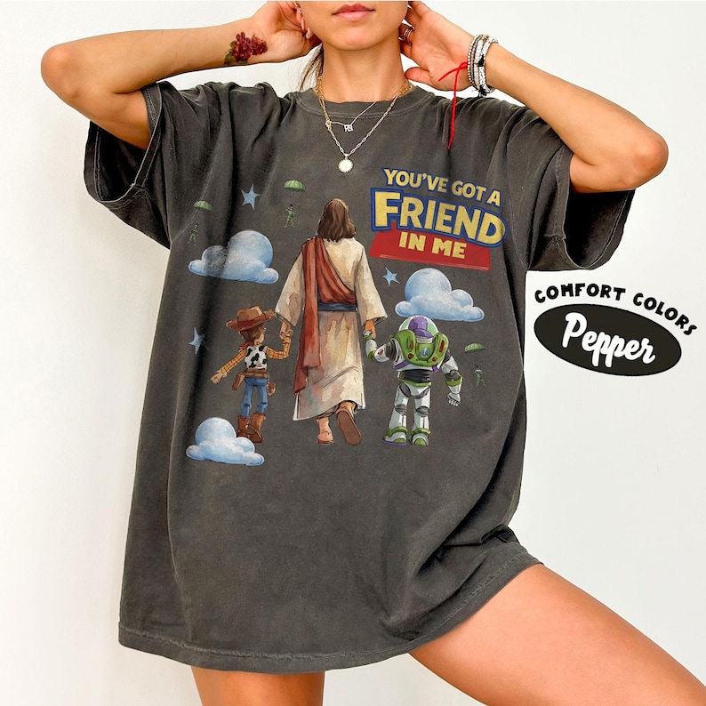 May include: Dark grey t-shirt featuring the text "YOU'VE GOT A FRIEND IN ME" above an illustration of Jesus, Woody, and Buzz Lightyear. The shirt also has the words "COMFORT COLORS" and "Pepper" in a white oval.