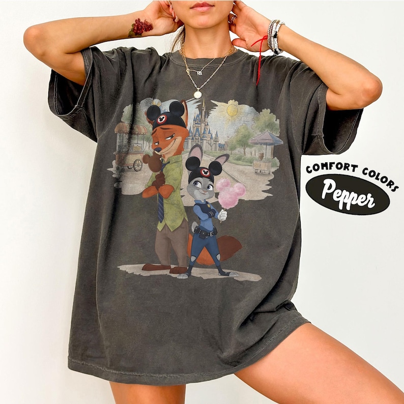 Puede incluir: Camiseta gris oscuro con una ilustraci&oacute;n de dibujos animados de Nick Wilde y Judy Hopps de Zootopia, con orejas de Mickey Mouse. La camiseta tiene el texto "COMFORT COLORS Pepper" en un &oacute;valo blanco.