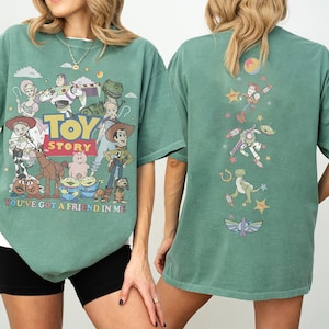 Op de afbeelding: Een saliegroen T-shirt met korte mouwen en een afbeelding van Toy Story-personages op de voorkant met de tekst TOY STORY en YOU'VE GOT A FRIEND IN ME. De achterkant van het shirt toont een verticale opstelling van personages en iconen.