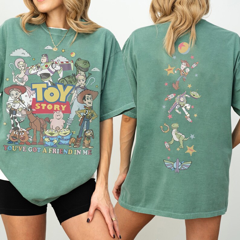 Op de afbeelding: Een saliegroen T-shirt met korte mouwen en een afbeelding van Toy Story-personages op de voorkant met de tekst TOY STORY en YOU'VE GOT A FRIEND IN ME. De achterkant van het shirt toont een verticale opstelling van personages en iconen.