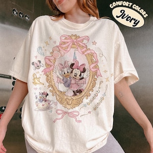 May include: Ivory-colored t-shirt featuring a framed illustration of Minnie and Daisy Duck in pink dresses. The shirt has a pink bow and the words "Disney Girls" in a cursive font. The shirt also has the words "Comfort Colors" and "Ivory" in a circular design.