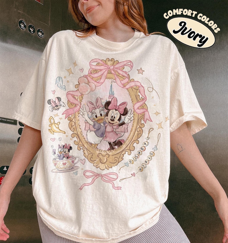 May include: Ivory-colored t-shirt featuring a framed illustration of Minnie and Daisy Duck in pink dresses. The shirt has a pink bow and the words "Disney Girls" in a cursive font. The shirt also has the words "Comfort Colors" and "Ivory" in a circular design.