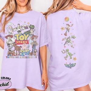Op de afbeelding: Een lichtpaars t-shirt met Toy Story-personages op de voor- en achterkant. De voorkant toont een groep personages met de tekst Toy Story en You've Got A Friend In Me. De achterkant toont een verticale rangschikking van personages en iconen.