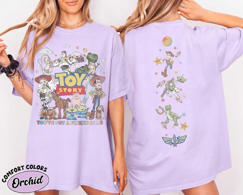Op de afbeelding: Een lichtpaars t-shirt met Toy Story-personages op de voor- en achterkant. De voorkant toont een groep personages met de tekst Toy Story en You've Got A Friend In Me. De achterkant toont een verticale rangschikking van personages en iconen.