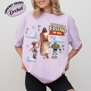 May include: A lilac t-shirt featuring the phrase "YOU'VE GOT A FRIEND IN ME" and a graphic of Jesus with Woody and Buzz Lightyear. The design includes clouds, stars, and parachutes. The shirt has the words "Comfort Colors" and "Orchid" in a circle.