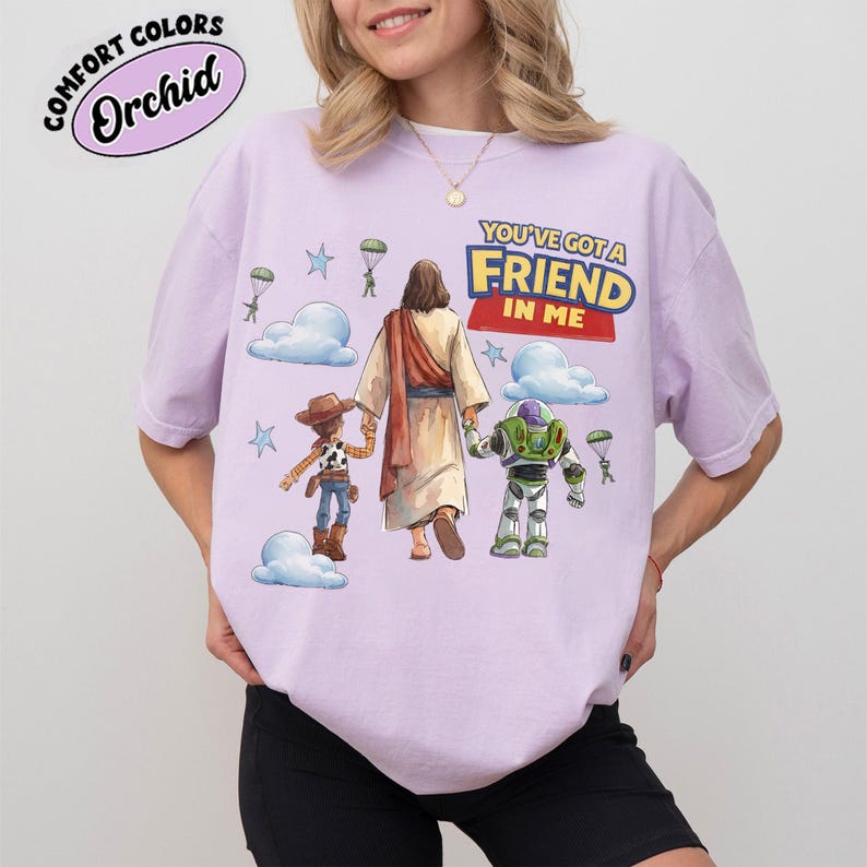 May include: A lilac t-shirt featuring the phrase "YOU'VE GOT A FRIEND IN ME" and a graphic of Jesus with Woody and Buzz Lightyear. The design includes clouds, stars, and parachutes. The shirt has the words "Comfort Colors" and "Orchid" in a circle.