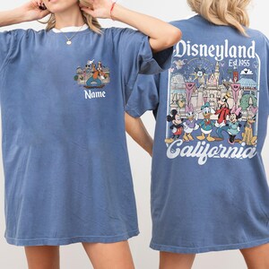 May include: Blue t-shirt with a white collar featuring a Disneyland graphic. The front has a cartoon image and the word "Name". The back displays a Disneyland scene with characters and the text "Disneyland Est. 1955 California".