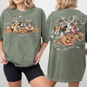 May include: Olive green t-shirt featuring a Halloween-themed graphic of Disney characters, including Mickey Mouse, Minnie Mouse, and Goofy, with carved pumpkins. The design is on the front and back of the shirt. The shirt has short sleeves and a relaxed fit.