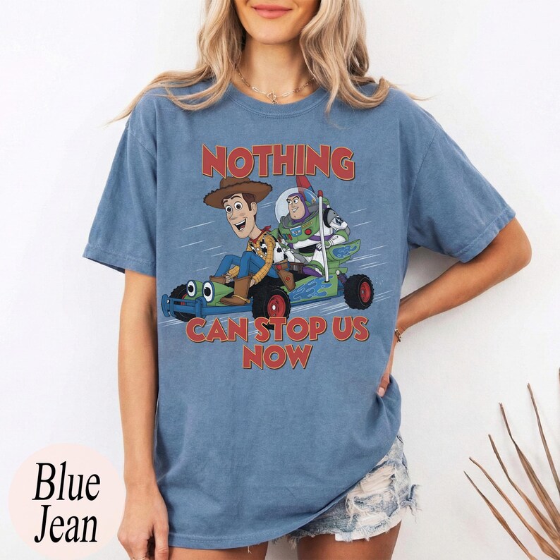 May include: A blue short-sleeve t-shirt featuring a graphic of Woody and Buzz Lightyear riding a go-kart. The text "NOTHING CAN STOP US NOW" is printed in red above and below the characters. The shirt has a crew neckline and a relaxed fit.