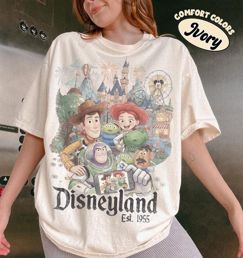 May include: An ivory short-sleeve t-shirt featuring a graphic print of Toy Story characters including Woody, Buzz Lightyear, Jessie, Rex, and Mr. Potato Head at Disneyland. The text reads Disneyland Est. 1955 and Comfort Colors Ivory.