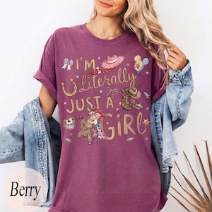 K&ouml;nnte beinhalten: Beerenfarbenes T-Shirt mit dem Text "I'm Literally Just a Girl" und Cartoon-Grafiken eines Cowboyhuts, Stiefeln und eines Pferdes. Das Shirt hat hochgekrempelte &Auml;rmel und wird mit einer Jeansjacke kombiniert.
