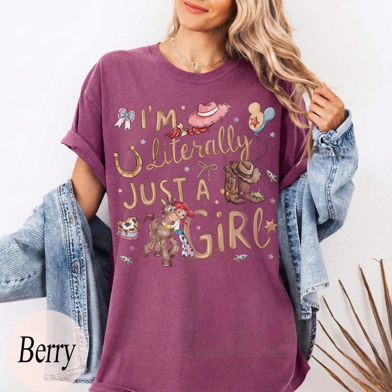 K&ouml;nnte beinhalten: Beerenfarbenes T-Shirt mit dem Text "I'm Literally Just a Girl" und Cartoon-Grafiken eines Cowboyhuts, Stiefeln und eines Pferdes. Das Shirt hat hochgekrempelte &Auml;rmel und wird mit einer Jeansjacke kombiniert.