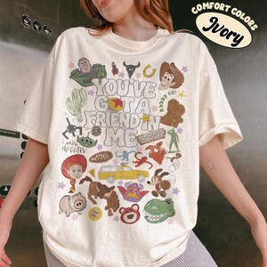 Op de afbeelding: Een ivoorkleurig T-shirt met korte mouwen en een grafische print met de tekst "YOU'VE GOT A FRIEND IN ME", omringd door diverse kleurrijke illustraties van stripfiguren, een gele vrachtwagen, een cactus en andere kleine iconen.