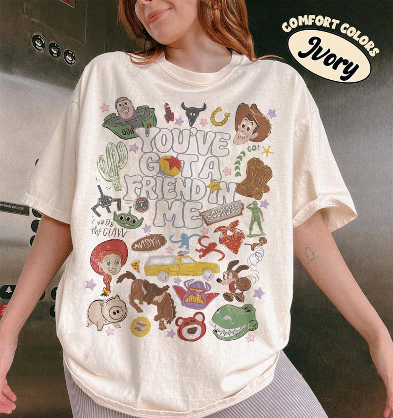 Op de afbeelding: Een ivoorkleurig T-shirt met korte mouwen en een grafische print met de tekst "YOU'VE GOT A FRIEND IN ME", omringd door diverse kleurrijke illustraties van stripfiguren, een gele vrachtwagen, een cactus en andere kleine iconen.