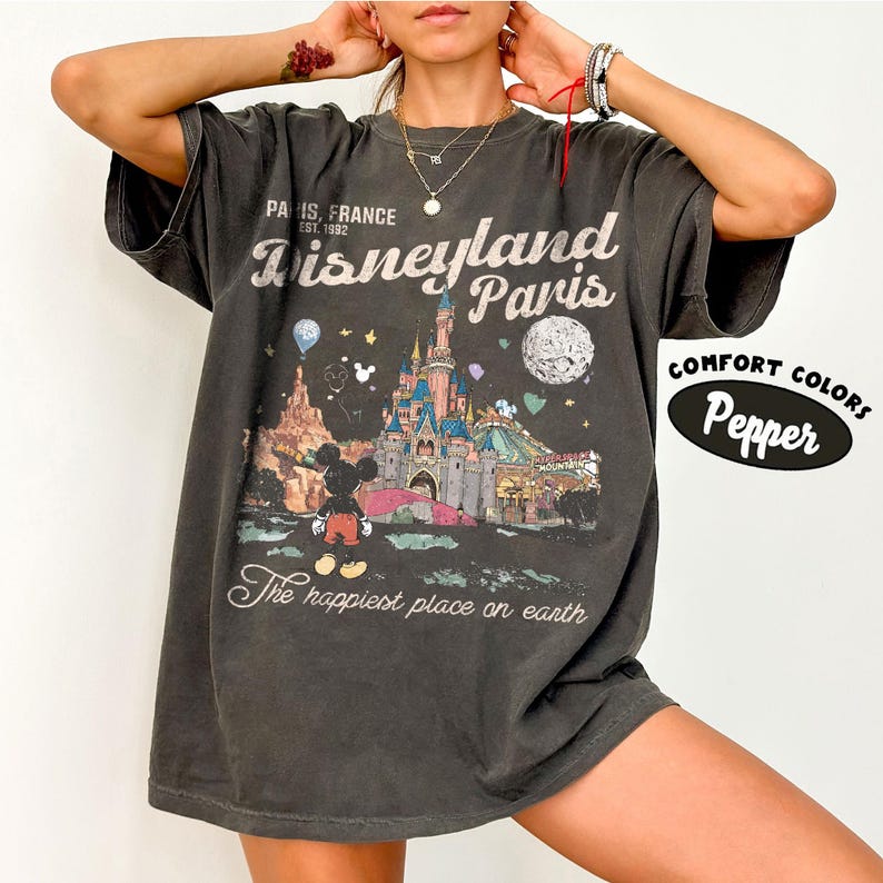 May include: Dark gray t-shirt featuring a Disneyland Paris graphic. The design includes the Disneyland Paris logo, a castle, Mickey Mouse, and the phrase "The happiest place on earth." The shirt has short sleeves and the words "Comfort Colors Pepper" on the right side.