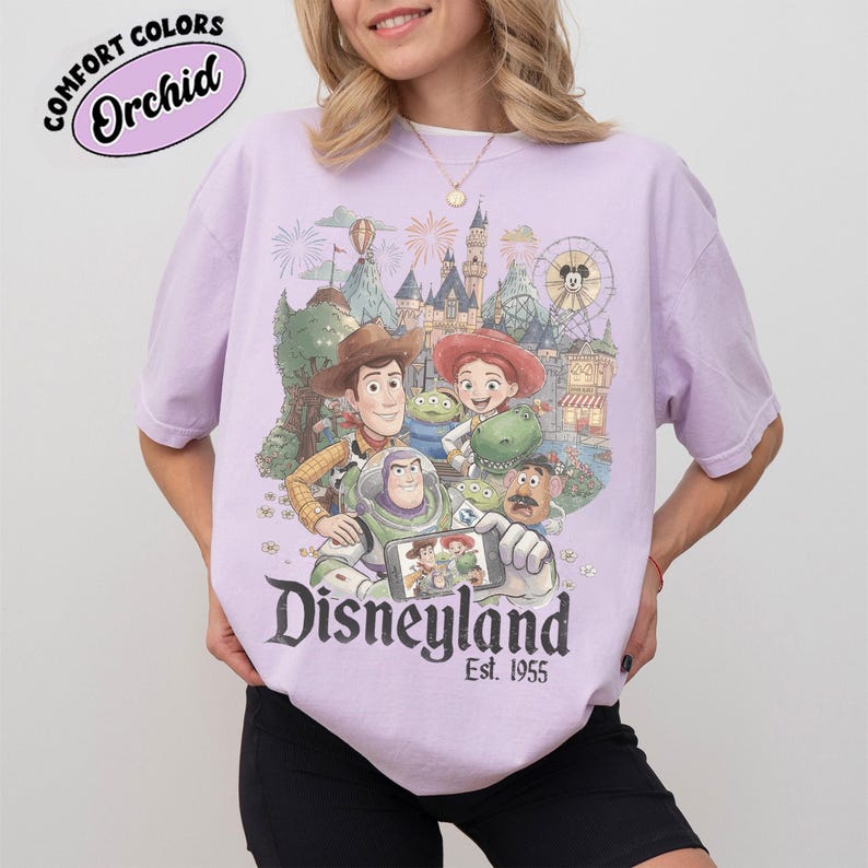 May include: A light purple t-shirt featuring a graphic print of Toy Story characters including Woody, Jessie, Buzz Lightyear, Rex, and Mr. Potato Head in front of a Disneyland castle. The text reads Disneyland Est. 1955 and Comfort Colors Orchid.