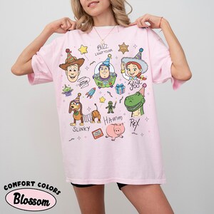 Pu&ograve; includere: T-shirt rosa chiaro con illustrazioni di personaggi dei cartoni animati di Toy Story. Il design include Woody, Buzz Lightyear, Jessie, Rex, Slinky, Hamm e altri personaggi.