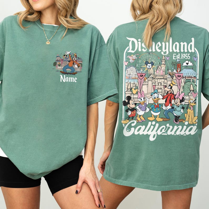 May include: A sage green t-shirt featuring a Disneyland graphic on the back and a smaller Goofy design on the front. The back graphic includes the Disneyland logo, the text "Est. 1955", and various cartoon characters. The front has the word "Name" below the Goofy design.
