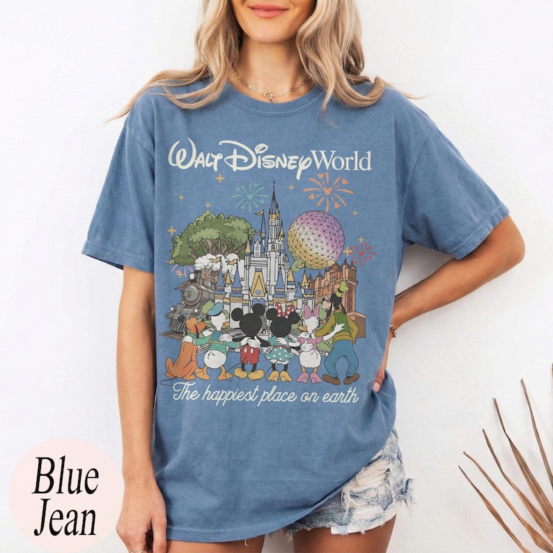 Puede incluir: Camiseta azul vaquera con un gr&aacute;fico de Walt Disney World. El dise&ntilde;o presenta el castillo de Disney World, personajes, fuegos artificiales y el texto "The happiest place on earth". La camiseta tambi&eacute;n tiene el texto "Walt Disney World" en la parte superior.