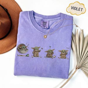 Puede incluir: Una camiseta violeta Comfort Colors con un gr&aacute;fico de un personaje de una popular franquicia de ciencia ficci&oacute;n. El personaje se representa en cuatro poses diferentes. Un collar dorado est&aacute; drapeado sobre la camiseta.