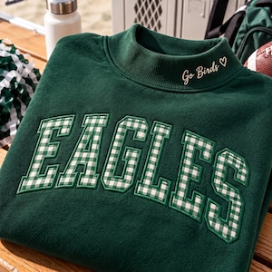 Op de afbeelding: Een donkergroene sweater met een opstaande kraag met de geborduurde tekst Go Birds en een hartsymbool. De voorkant toont het woord EAGLES in grote blokletters met een wit en groen gingham ruitpatroon.