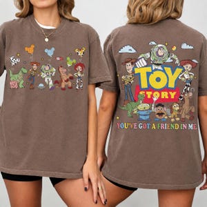 May include: A brown t-shirt featuring Toy Story characters. The front displays small illustrations of Woody, Buzz Lightyear, Rex, and Jessie with balloons. The back shows a large Toy Story logo with the characters and the text You've Got A Friend In Me.