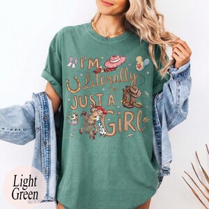 K&ouml;nnte beinhalten: Hellgr&uuml;nes T-Shirt mit dem Text "I'm Literally Just a Girl" und Cowboy-Grafiken. Das Design umfasst ein Hufeisen, Cowboystiefel, einen Hut und ein Cartoon-Pferd mit einem Cowgirl. Das Shirt hat eine lockere Passform.