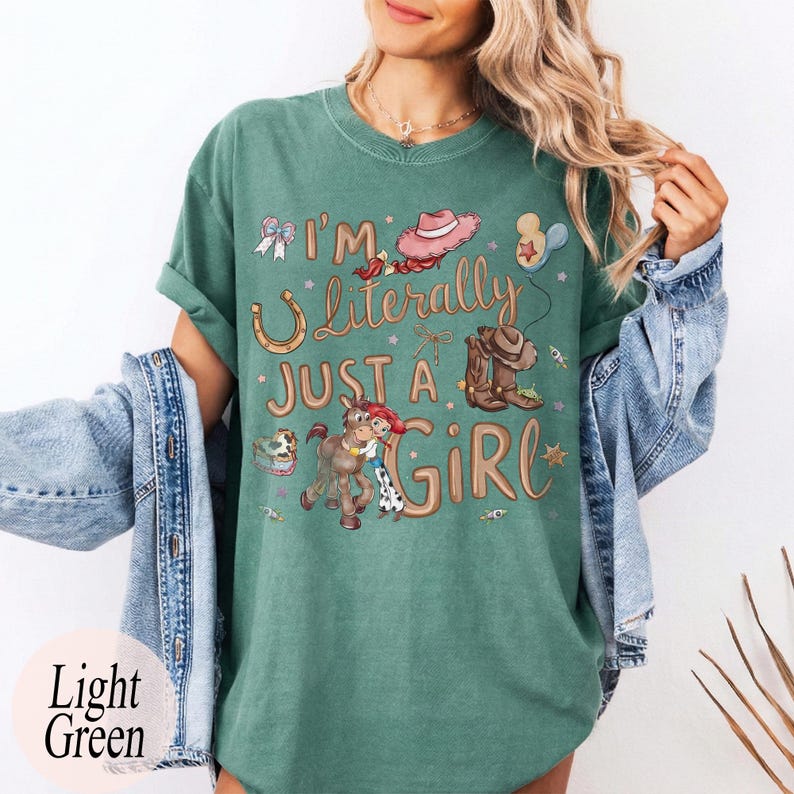 K&ouml;nnte beinhalten: Hellgr&uuml;nes T-Shirt mit dem Text "I'm Literally Just a Girl" und Cowboy-Grafiken. Das Design umfasst ein Hufeisen, Cowboystiefel, einen Hut und ein Cartoon-Pferd mit einem Cowgirl. Das Shirt hat eine lockere Passform.