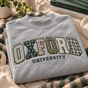 May include: A light grey crewneck sweatshirt featuring the word OXFORD in large, multi-patterned fabric letters with various designs including stripes, stars, hearts, and gingham checks, with the word UNIVERSITY embroidered in dark green below.
