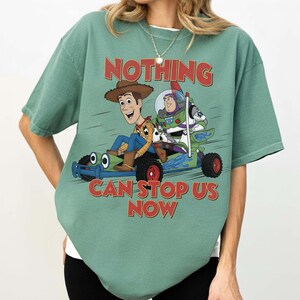 May include: A sage green short-sleeve t-shirt featuring a graphic of Woody and Buzz Lightyear riding a go-kart. The text "NOTHING CAN STOP US NOW" is printed in red above and below the characters. The shirt has a crew neckline and a loose fit.