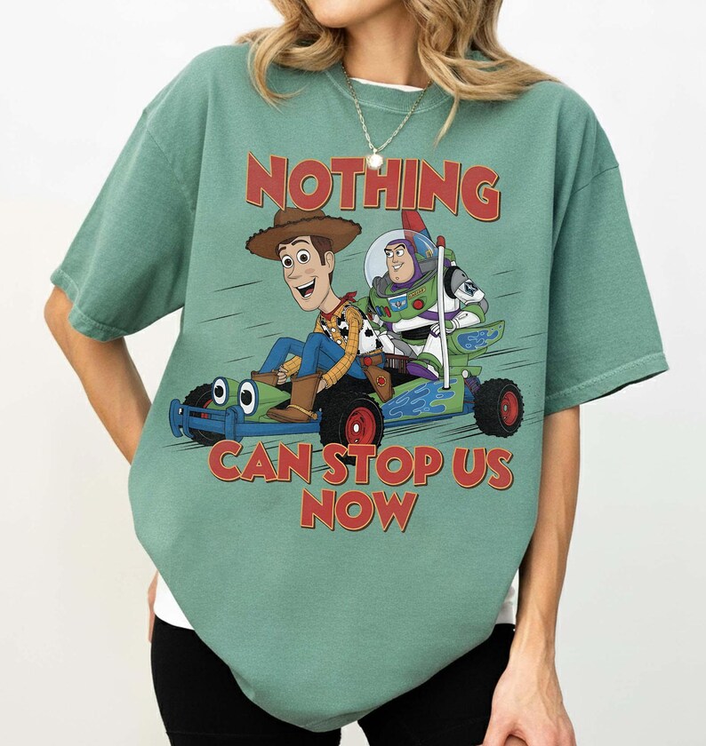 May include: A sage green short-sleeve t-shirt featuring a graphic of Woody and Buzz Lightyear riding a go-kart. The text "NOTHING CAN STOP US NOW" is printed in red above and below the characters. The shirt has a crew neckline and a loose fit.