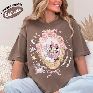 May include: Espresso-colored t-shirt featuring a framed illustration of Minnie and Daisy Duck. The design includes pink bows, stars, and other decorative elements. The shirt also has the text "COMFORT COLORS" and "Espresso" in a circular design.