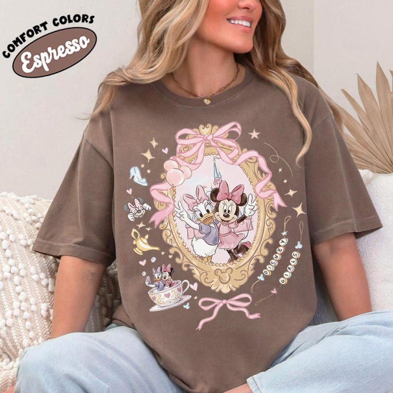 May include: Espresso-colored t-shirt featuring a framed illustration of Minnie and Daisy Duck. The design includes pink bows, stars, and other decorative elements. The shirt also has the text "COMFORT COLORS" and "Espresso" in a circular design.