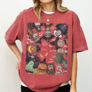 May include: A brick red short-sleeve t-shirt featuring a rectangular graphic print of various animated characters in Halloween costumes. The design includes labels for characters like Woody, Buzz Lightyear, Jessie, Rex, Forky, Ham, and others, surrounded by small white bats.