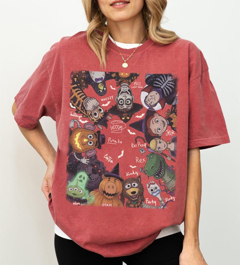 May include: A brick red short-sleeve t-shirt featuring a rectangular graphic print of various animated characters in Halloween costumes. The design includes labels for characters like Woody, Buzz Lightyear, Jessie, Rex, Forky, Ham, and others, surrounded by small white bats.