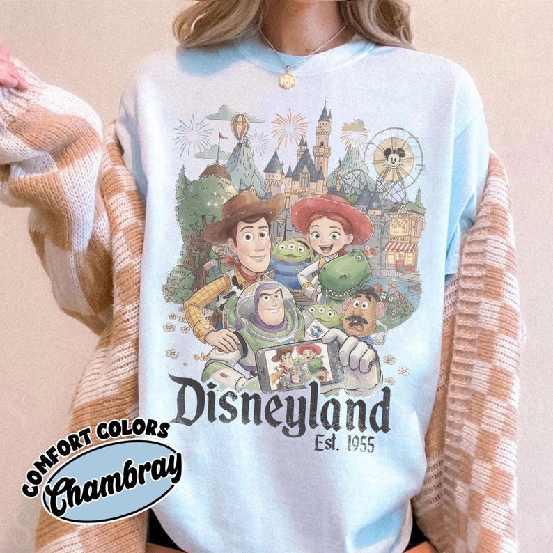 May include: A light blue crewneck sweatshirt featuring a graphic print of various animated characters, including a cowboy, a space ranger, and a cowgirl, in front of a theme park castle. The text Disneyland Est. 1955 is printed below the characters.