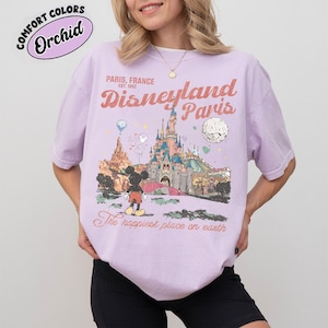 May include: A light purple t-shirt with a Disneyland Paris graphic. The graphic features a castle, Mickey Mouse, and the text "Paris, France, Disneyland Paris, The happiest place on earth." The shirt has short sleeves and a crew neck.
