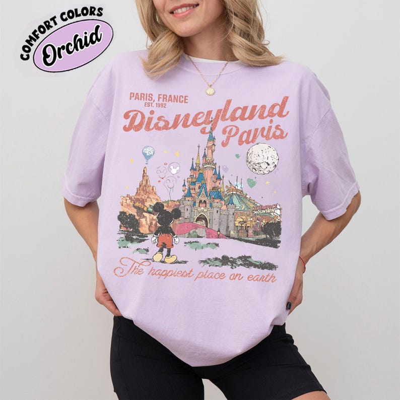 May include: A light purple t-shirt with a Disneyland Paris graphic. The graphic features a castle, Mickey Mouse, and the text "Paris, France, Disneyland Paris, The happiest place on earth." The shirt has short sleeves and a crew neck.