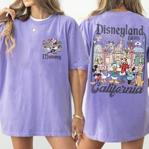 May include: A light purple t-shirt featuring a Minnie Mouse graphic with the word "Mommy" below. The back of the shirt displays a Disneyland graphic with various Disney characters and the text "Disneyland Est. 1955 California".