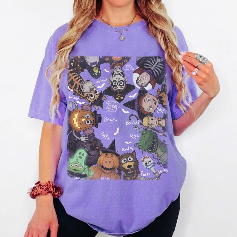 May include: A light purple short-sleeve t-shirt featuring a graphic print of various animated characters in Halloween costumes. The design includes labels for characters like Woody, Buzz Lightyear, Jessie, Bo Peep, Rex, Forky, Slinky, Hamm, and Alien.