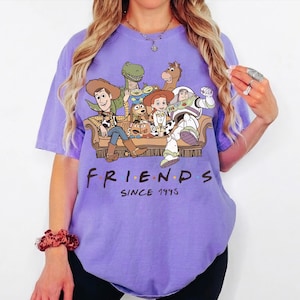 May include: A light purple short-sleeve t-shirt featuring a graphic print of various animated characters sitting on a couch. Below the illustration, the text reads FRIENDS SINCE 1995 in a bold, dark font. The shirt has a crew neckline and a relaxed fit.