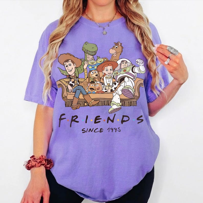 May include: A light purple short-sleeve t-shirt featuring a graphic print of various animated characters sitting on a couch. Below the illustration, the text reads FRIENDS SINCE 1995 in a bold, dark font. The shirt has a crew neckline and a relaxed fit.