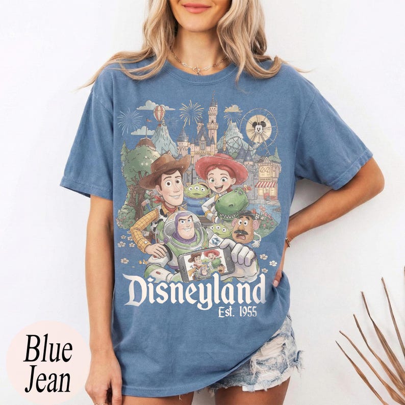 May include: A blue short-sleeve t-shirt featuring a graphic print of Toy Story characters taking a selfie in front of a Disneyland castle. The text Disneyland Est. 1955 is printed below the characters in a white serif font on the front of the shirt.