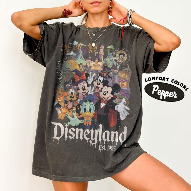 May include: A charcoal gray short-sleeve t-shirt featuring a graphic of Disney characters in Halloween costumes in front of a castle. The text reads Disneyland Est. 1955. A separate oval graphic on the right side displays the text Comfort Colors Pepper.