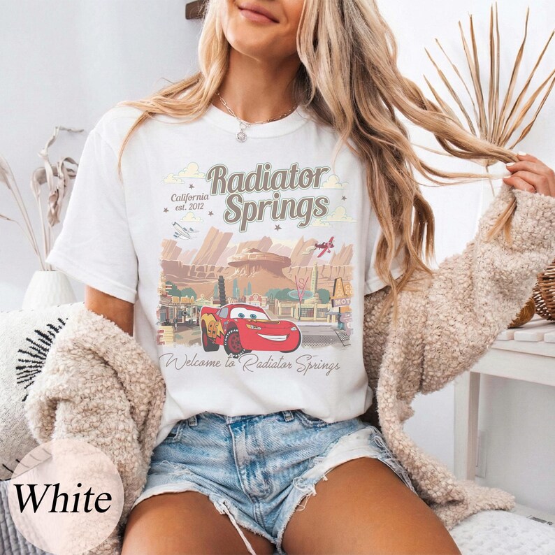 May include: White t-shirt with a graphic of Radiator Springs from the film Cars. The design includes the text "Radiator Springs" and "Welcome to Radiator Springs". The shirt also features a graphic of Lightning McQueen.