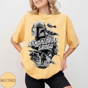 Op de afbeelding: Mosterdgeel T-shirt met een zwart-witte afbeelding uit de Star Wars-serie, "The Mandalorian and Grogu". Het ontwerp bevat de helm van de Mandalorian, Grogu en de zin "This is the Way."