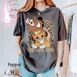 May include: Dark gray t-shirt featuring a graphic of characters from The Lion King, including Simba wearing a crown, Pumbaa, and Timon. The text "THE LION KING" is printed below the characters. The shirt has a relaxed fit.